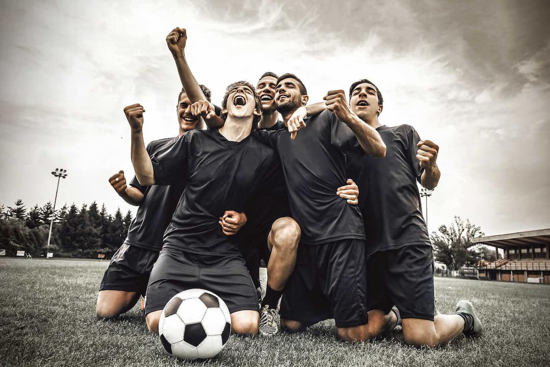 A soccer team celebrating on their needs in the middles of a field with a soccer ball in front of them
